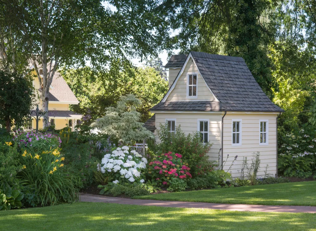 A small backyard cottage painted a light yellow color surrounded by white, yellow, and purple flowers.