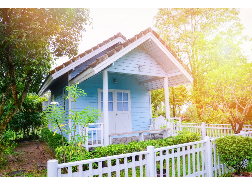 A small backyard cottage painted a light blue with a white picket fence surrounding it.
